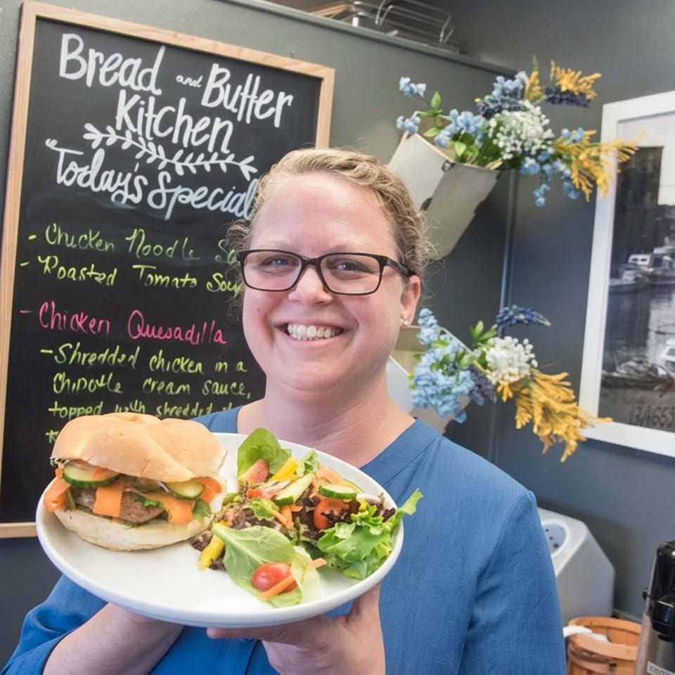 Smiling woman wearing glasses holds a plate with a sandwich and fresh salad inside Bread and Butter Kitchen.
