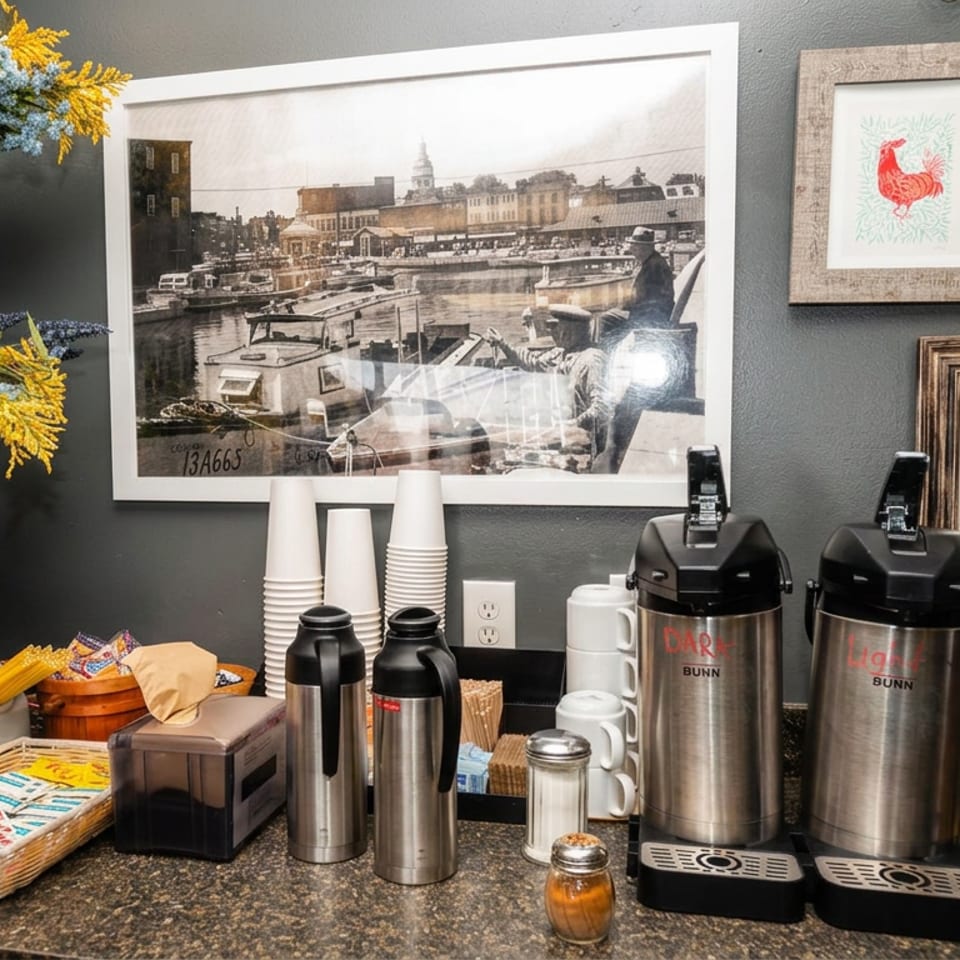 Coffee station with insulated dispensers, cups, condiments, snack baskets, floral wall décor, and framed artwork.