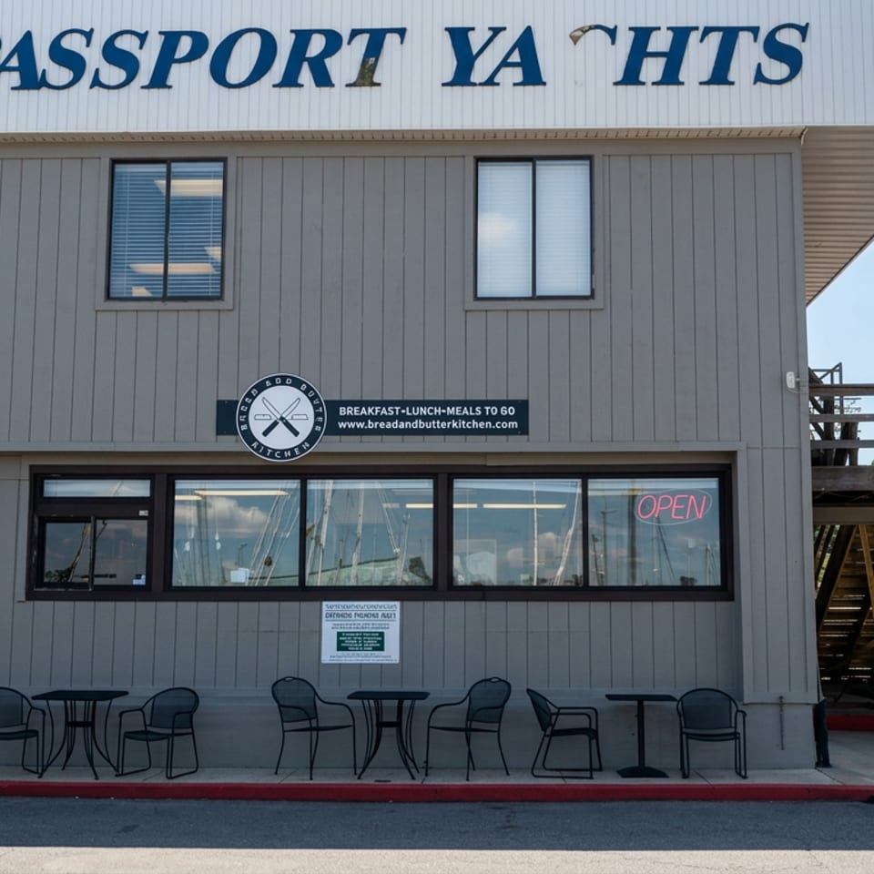 Exterior of Passport Yacht restaurant featuring outdoor tables, Bread and Butter Kitchen signage, open window, and a person walking nearby.
