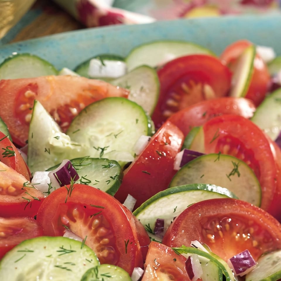 Close-up of a fresh tomato and cucumber salad with red onion and dill in a blue bowl.