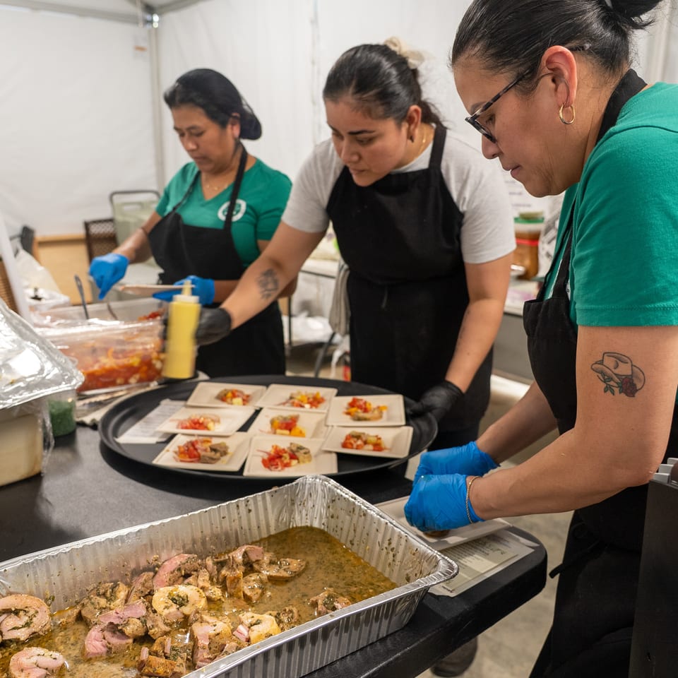 Caterers in aprons and gloves assemble plated dishes beside trays of sliced meat at a busy food-service station.