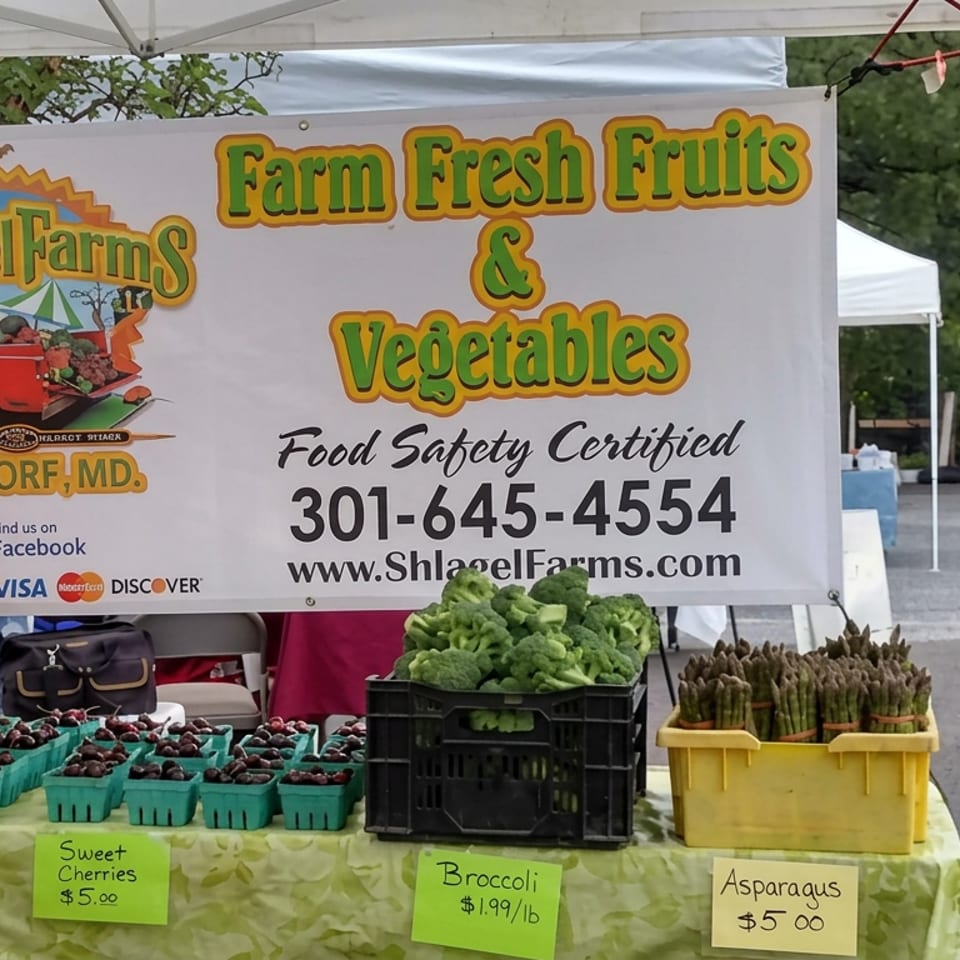 Shagel Farms market stall displays cherries, broccoli, and asparagus beneath a sign advertising farm-fresh fruits and vegetables in Waldorf, Maryland.