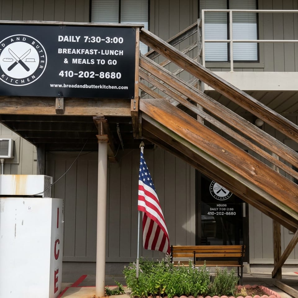 Exterior entrance of Bread and Butter Kitchen with wooden stairs, restaurant signage, American flag, bench, and planted flowers.